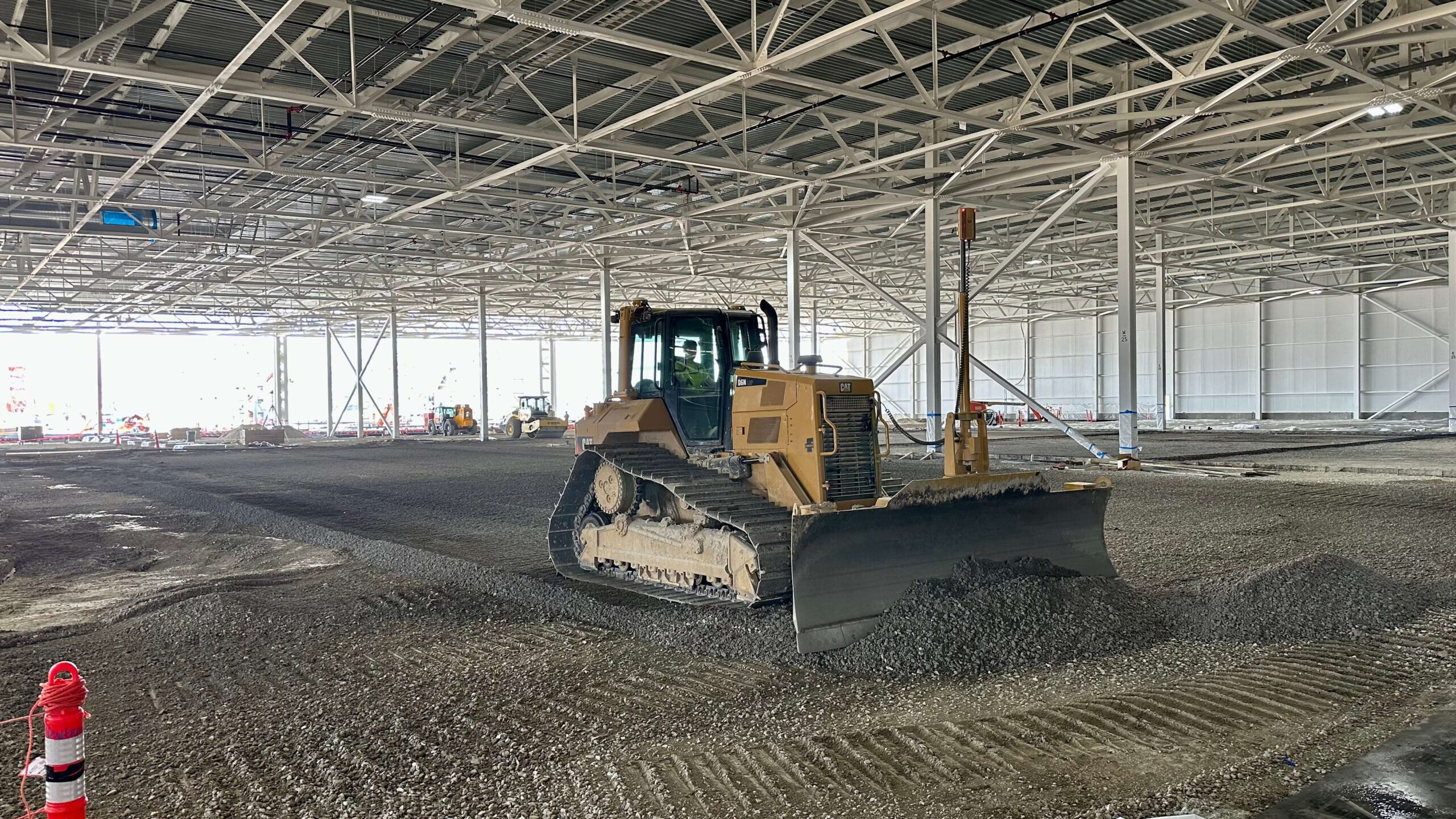 A bulldozer levels gravel inside a large industrial building under construction at Blue Oval City. The interior, marked by metal beams and a high ceiling, echoes the ambitious vision of Ford. Construction materials are scattered around, with a red safety cone visible in the foreground.