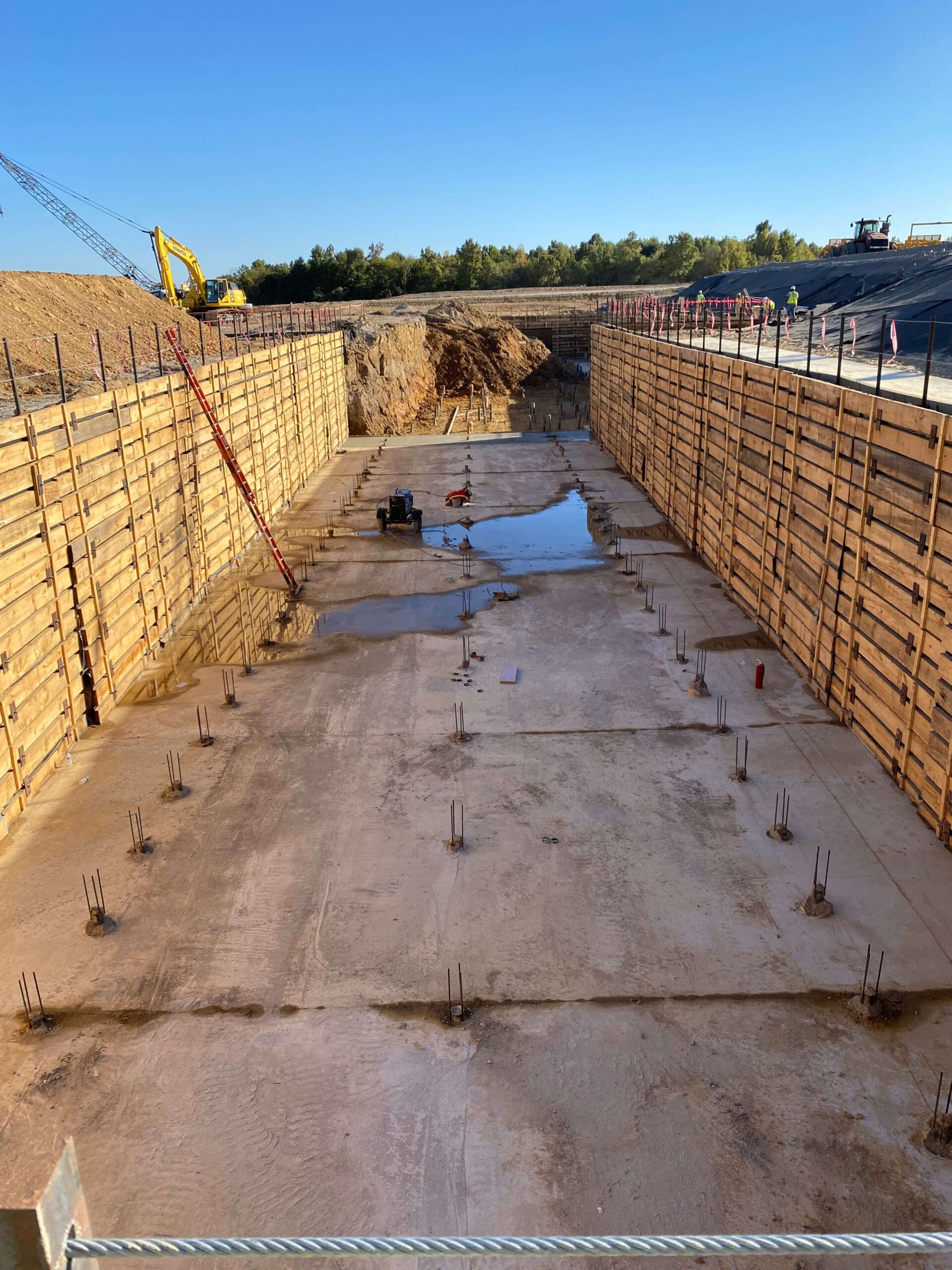 A construction site at the future Blue Oval City showcases a large, rectangular concrete foundation surrounded by wooden formwork. Reinforcement bars jut out as a crane looms in the background. A worker stands near a puddle on the foundation under the expansive blue sky.