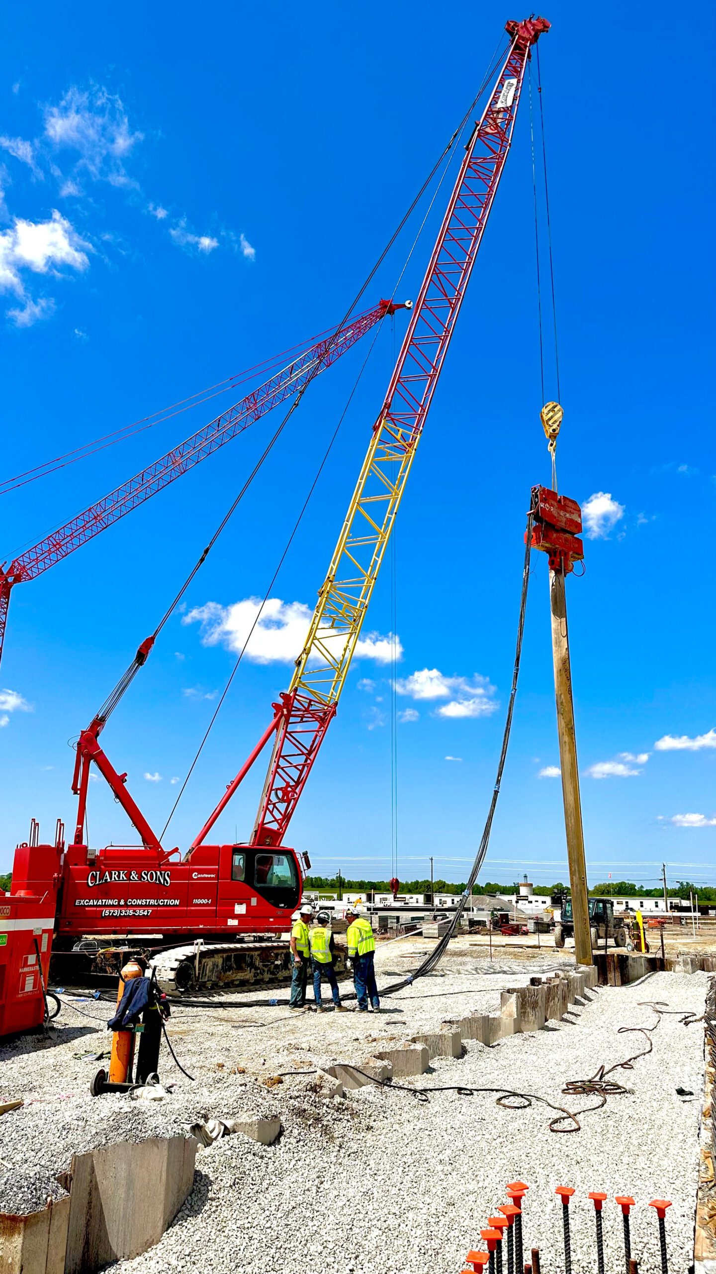 Red and yellow crane lifting large wooden pole at a construction site, with workers observing.