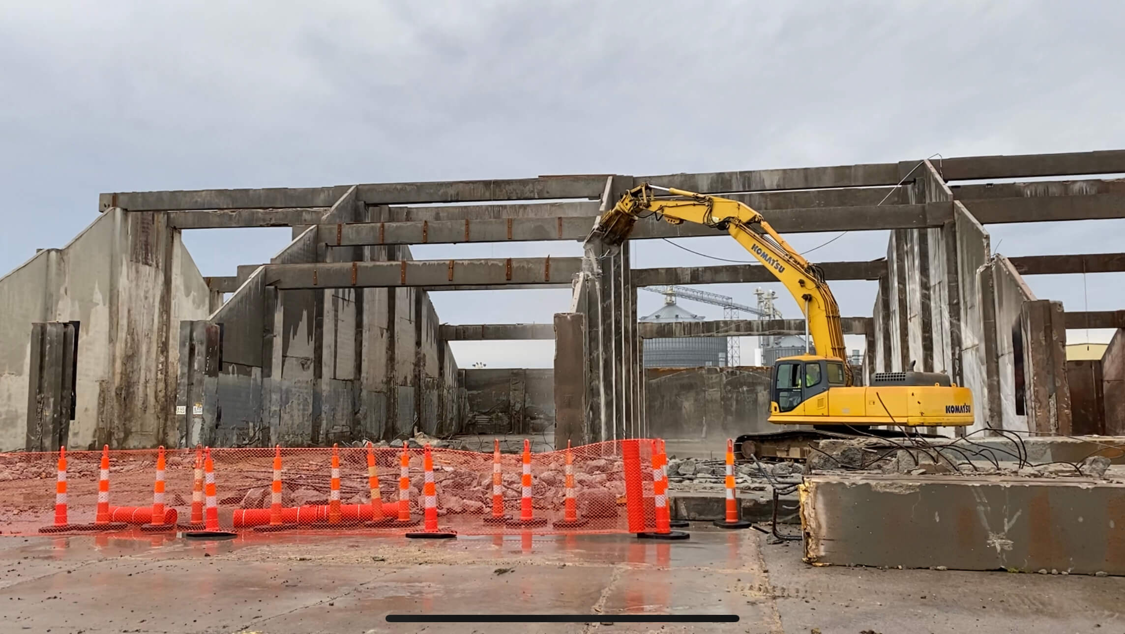 A yellow excavator, much like those used by Bruce Oakley, demolishes an industrial building, exposing large concrete beams. Orange traffic cones line the site in front of a pile of debris under an overcast sky.