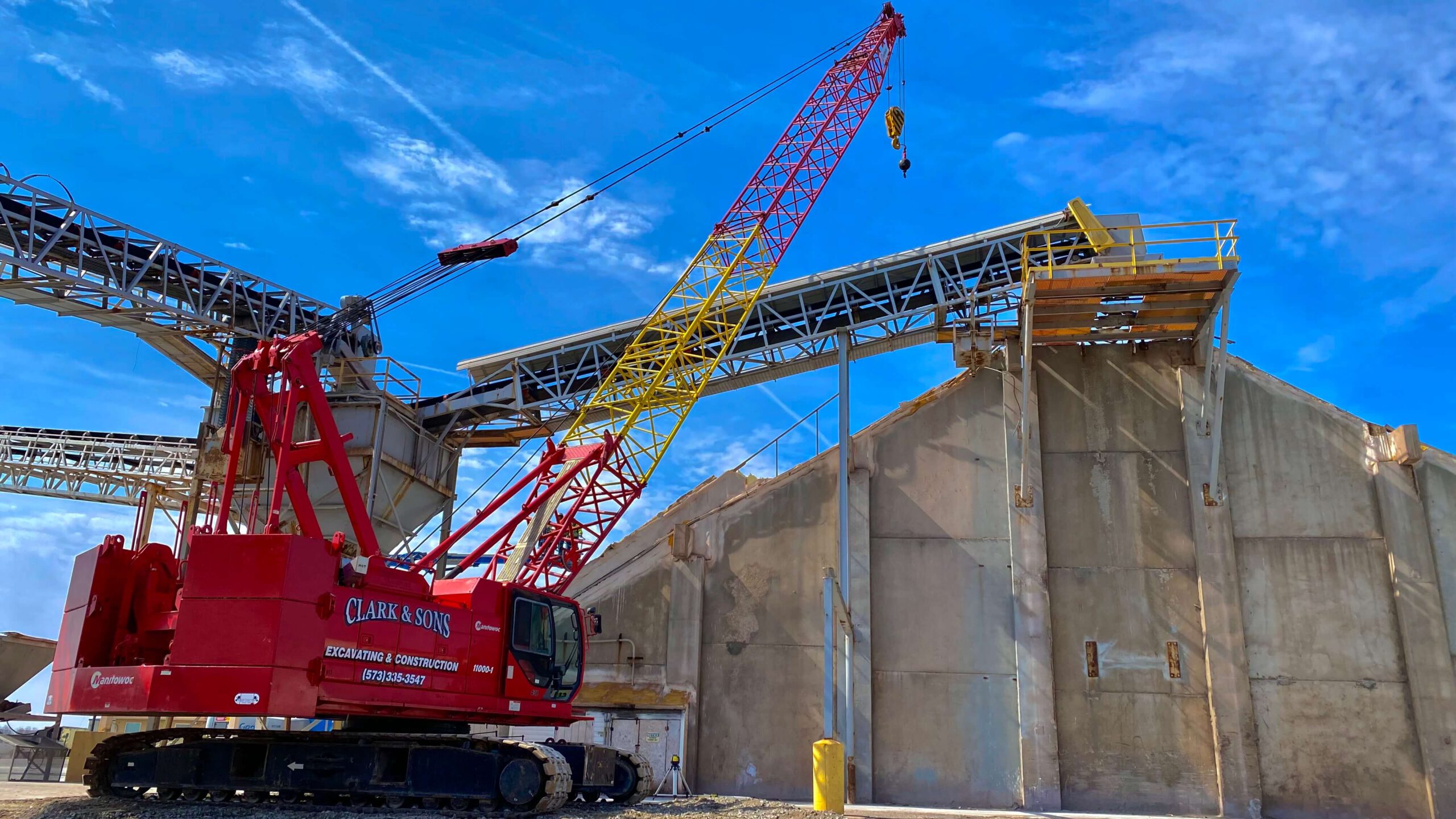 A red construction crane with a yellow boom, branded Clark & Sons, is positioned beside a large concrete industrial building under a blue sky. The site, reminiscent of Bruce Oakley projects, showcases metal frameworks and intricate structures surrounding the area.