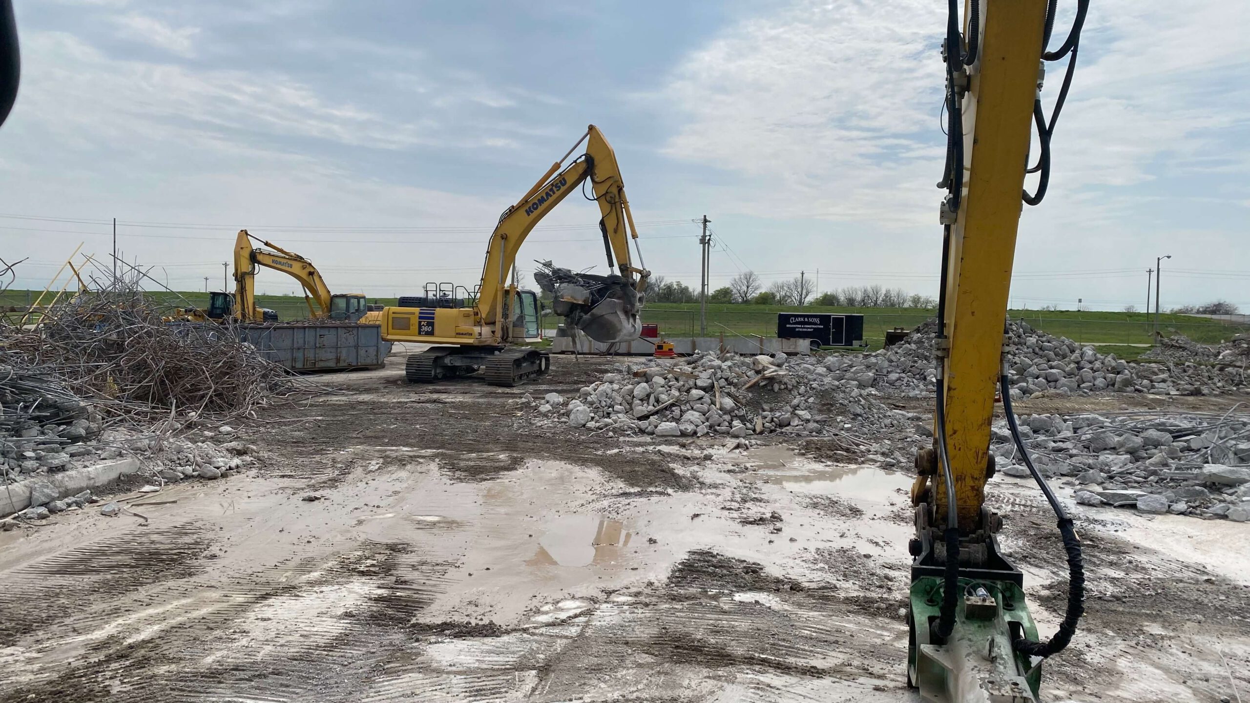 A construction site bustling with several Bruce Oakley excavators tirelessly working amidst piles of rubble and debris. The overcast sky casts a somber tone, while puddles dot the muddy ground. In the distance, fields and power lines quietly observe the scene.