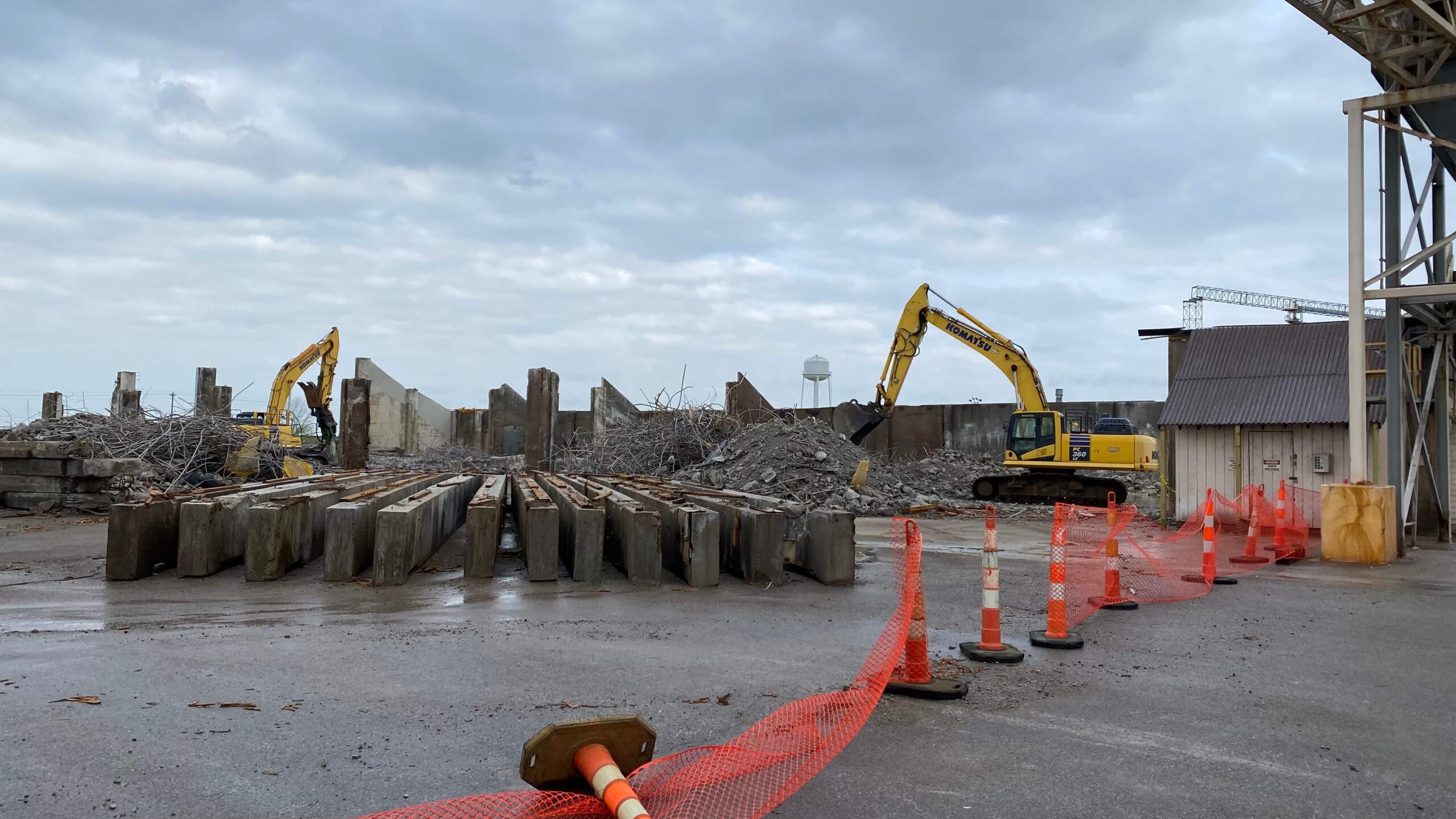 A construction site with two yellow excavators from Bruce Oakley stands amid rubble piles and large concrete beams. Orange fencing and traffic cones dot the foreground, all under an overcast sky.