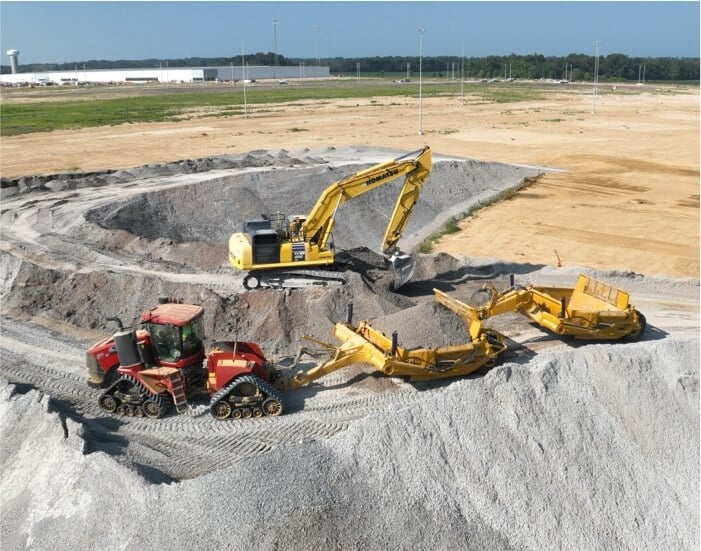 Construction site with three heavy machinery vehicles working on gravel piles. A yellow excavator and two other vehicles are moving earth, against a background of open land.